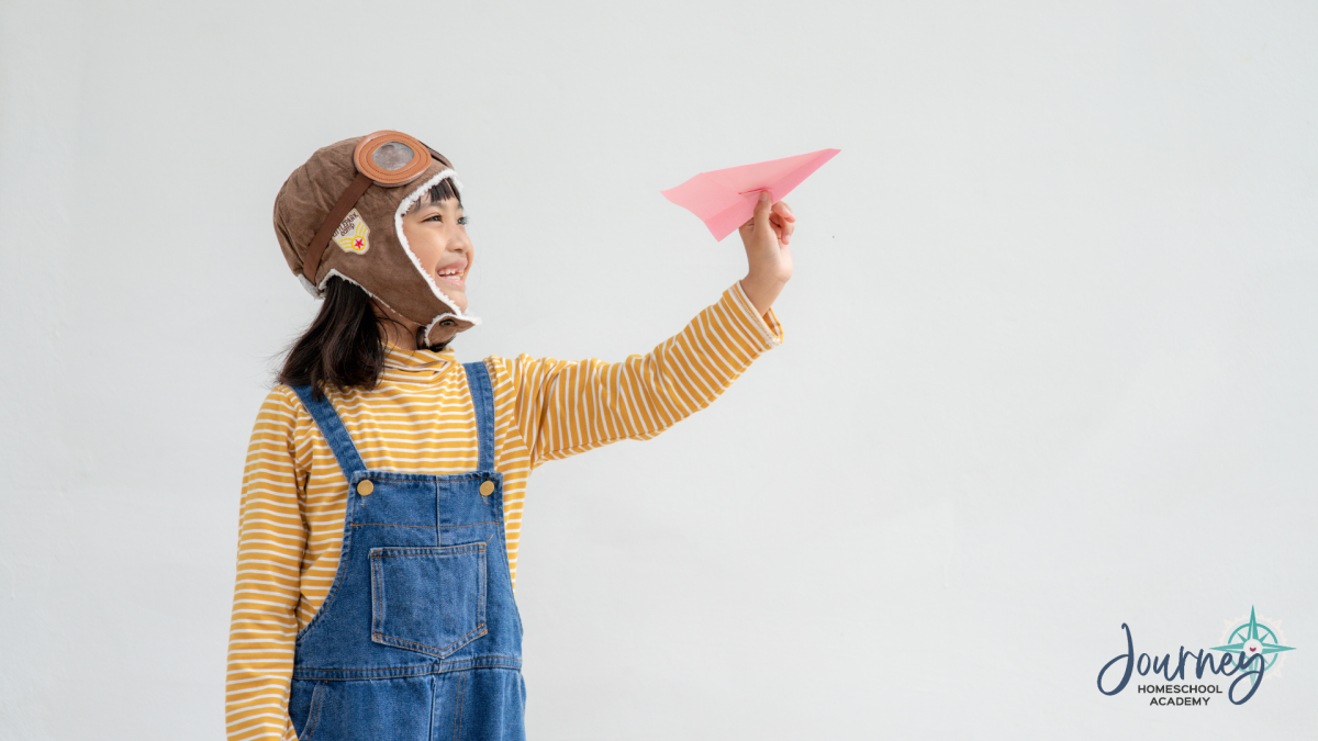 Child dressed as an aviator holding a paper airplane, illustrating homeschool physics of flight with Journey Homeschool Academy.