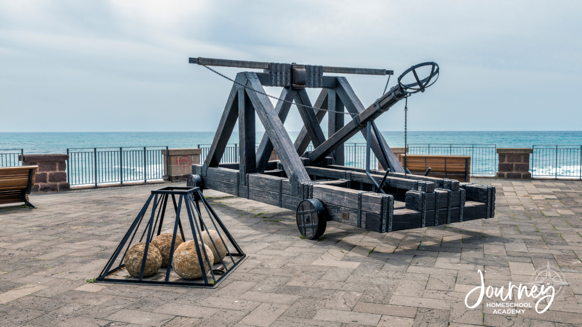 Full-sized reconstructed medieval catapult positioned on a seaside promenade with stone balls nearby, illustrating how ancient physics principles connect to modern engineering.