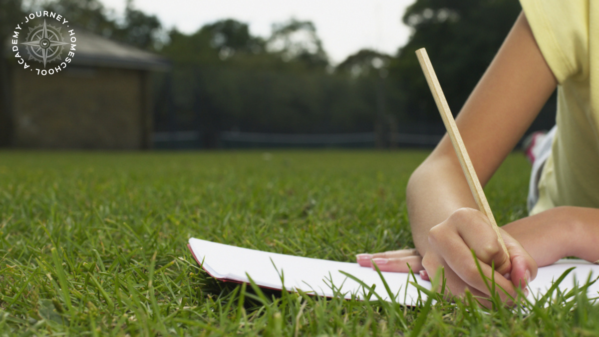 Child writing in a nature journal while lying in the grass, documenting a bird nesting homeschool project with Journey Homeschool Academy.