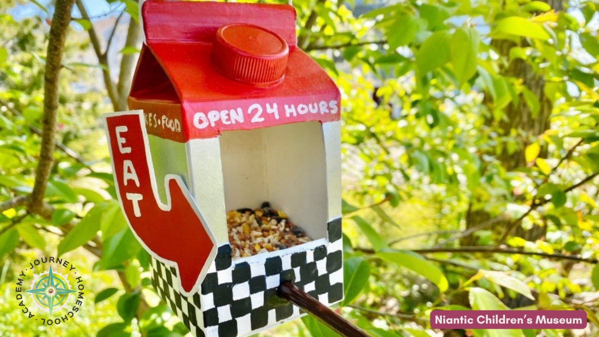 Painted milk carton bird feeder hanging outdoors with a retro “Open 24 Hours” sign, an example of a homeschool-friendly DIY bird feeder craft.
